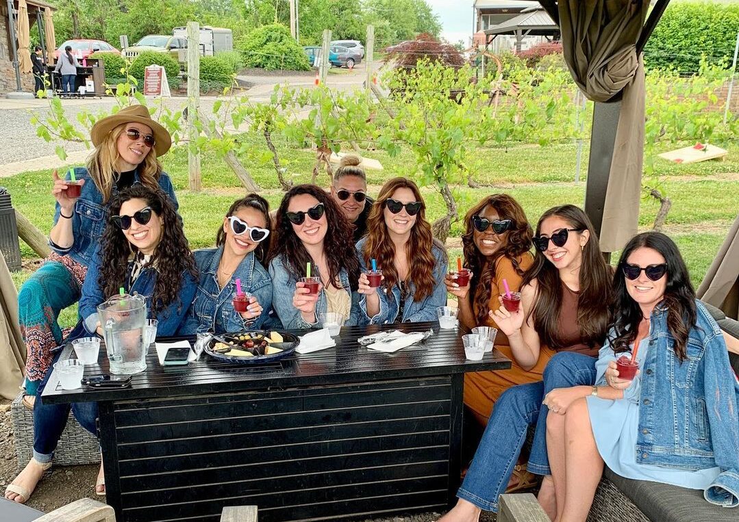 Group of nine women seated outdoors at Old York Cellars, smiling and holding glasses of red wine around a table under a private wine cabana.