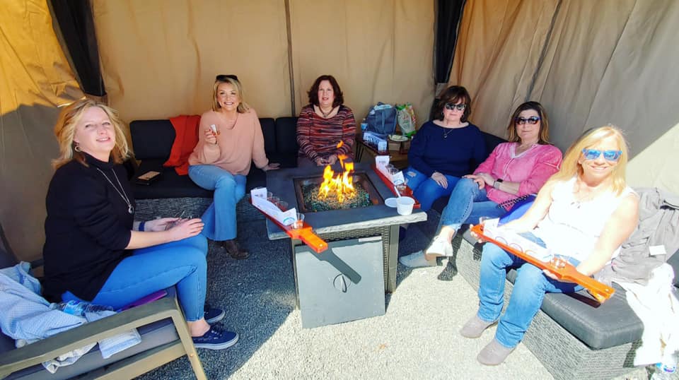 Six women sit around a rectangular fire pit inside a cozy outdoor tent at Old York Cellars. They are smiling and chatting, enjoying a relaxed afternoon together. Two hold long orange items, and the fire glows warmly in the center.