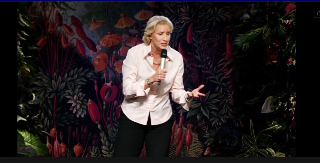 Helene Angley performing on stage in front of a colorful backdrop, holding a microphone and engaging the audience.  