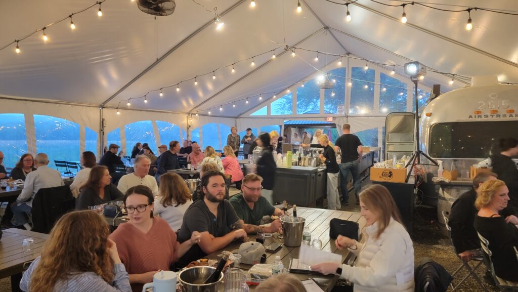 Guests enjoying a wine dinner at Old York Cellars before the start of the Wine & Comedy Show in Ringoes, Central NJ, seated under a vineyard tent with string lights and white‑clothed tables filled with wine glasses and plated courses.