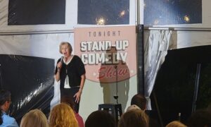 Comedian Helene Angley performs on stage at Old York Cellars during a Wine & Comedy Night. She stands before a banner reading “Tonight Stand‑Up Comedy Show,” holding a microphone and engaging the seated audience inside the winery’s tented venue in Ringoes, NJ