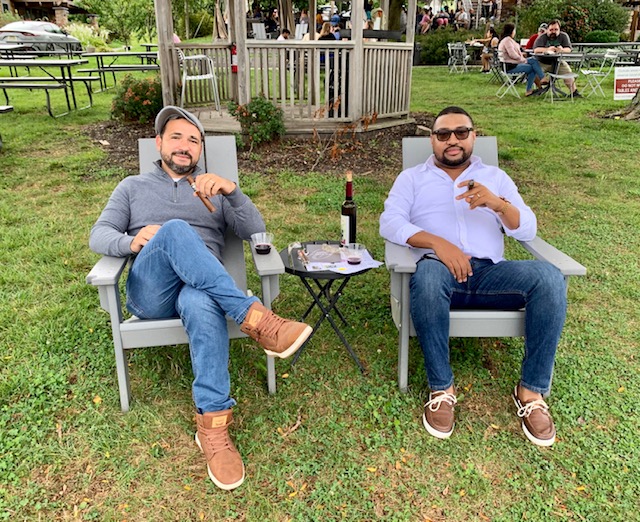 Two guests relaxing in outdoor chairs at Old York Cellars with a bottle of wine and glasses on a small table, surrounded by greenery and other visitors.
