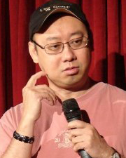 Comedian Bill Chiang in a pink shirt and black cap holding a microphone, performing on stage in front of a red curtain.