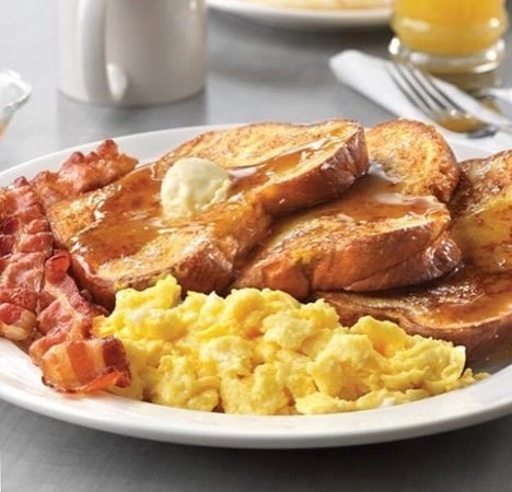 Plate of scrambled eggs, bacon, and French toast topped with butter and syrup on a table.