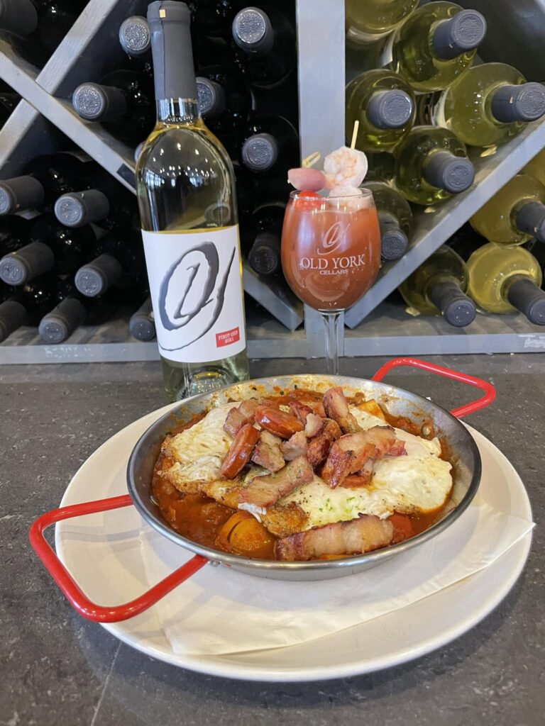 Huevos brunch dish served at Old York Cellars Winery in Ringoes, Hunterdon County, NJ, featuring eggs, potatoes, and sliced meat in a red‑handled pan on a white plate, paired with a bottle of Old York Cellars white wine and a Bloody Mary wine cocktail, displayed in front of a wine rack.