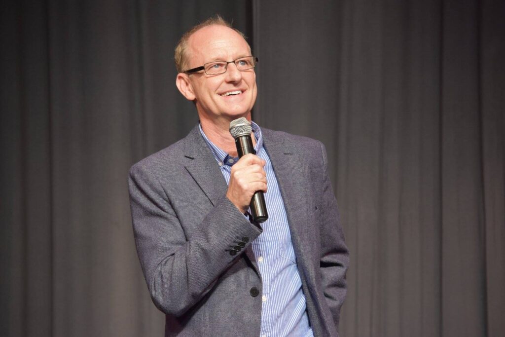 Comedian Buddy Fitzpatrick holding a microphone and smiling on stage, wearing glasses and a light jacket.  