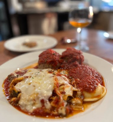 A plate of pasta topped with melted cheese and tomato sauce, served with two meatballs and a glass of wine in the background at a restaurant serving Old York Cellars wines.