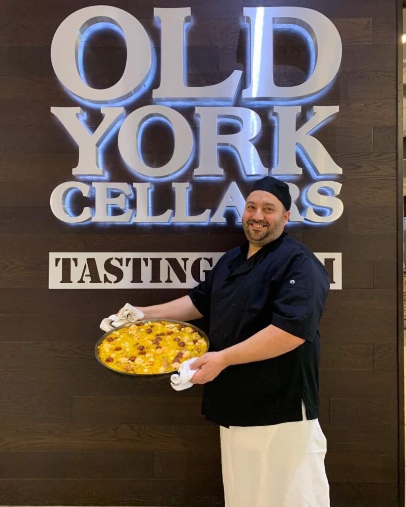 Chef José Diaz smiling indoors at Old York Cellars Tasting Room, holding a large pan of paella in front of wall with the winery’s logo.