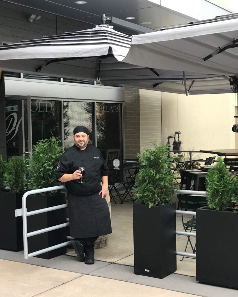 Chef José Diaz standing outside the Old York Cellars restaurant entrance at their former Quakerbridge Mall location, wearing a black chef’s coat and hat beside tall planters under a patio canopy.