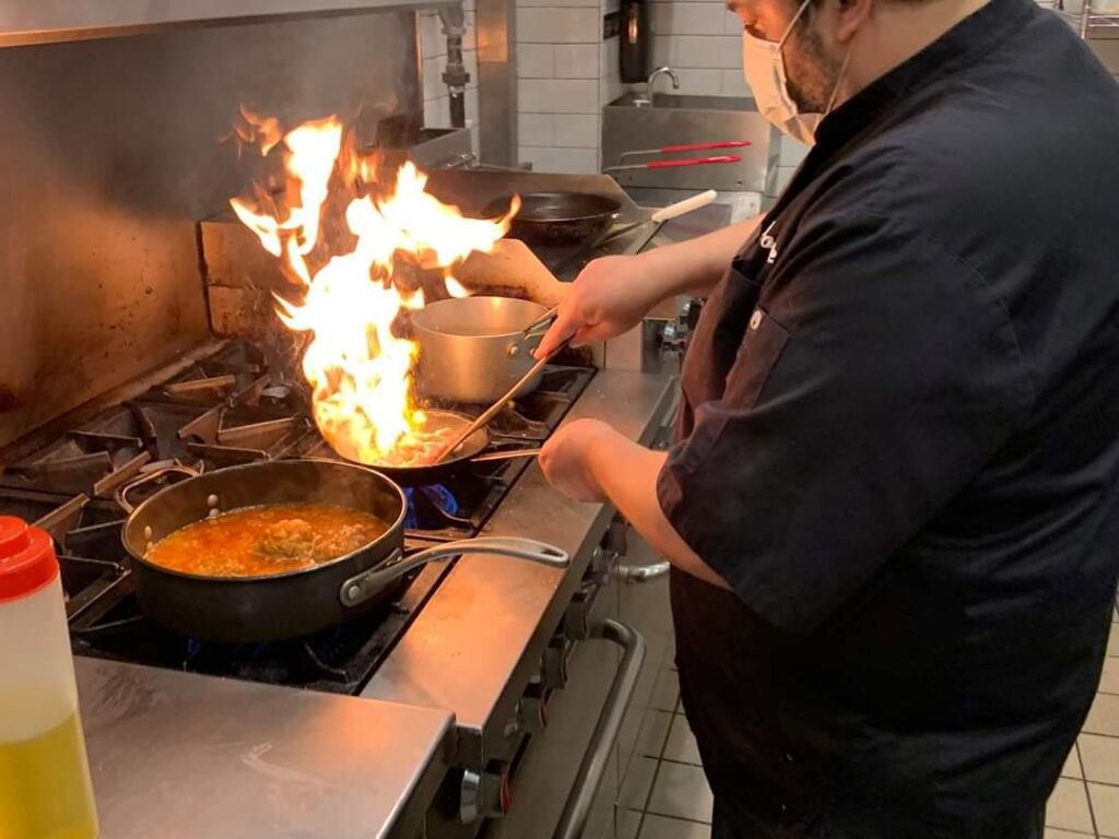 Chef José Diaz cooking in a professional kitchen, stirring pans on a stove with visible flames rising during high‑heat preparation.