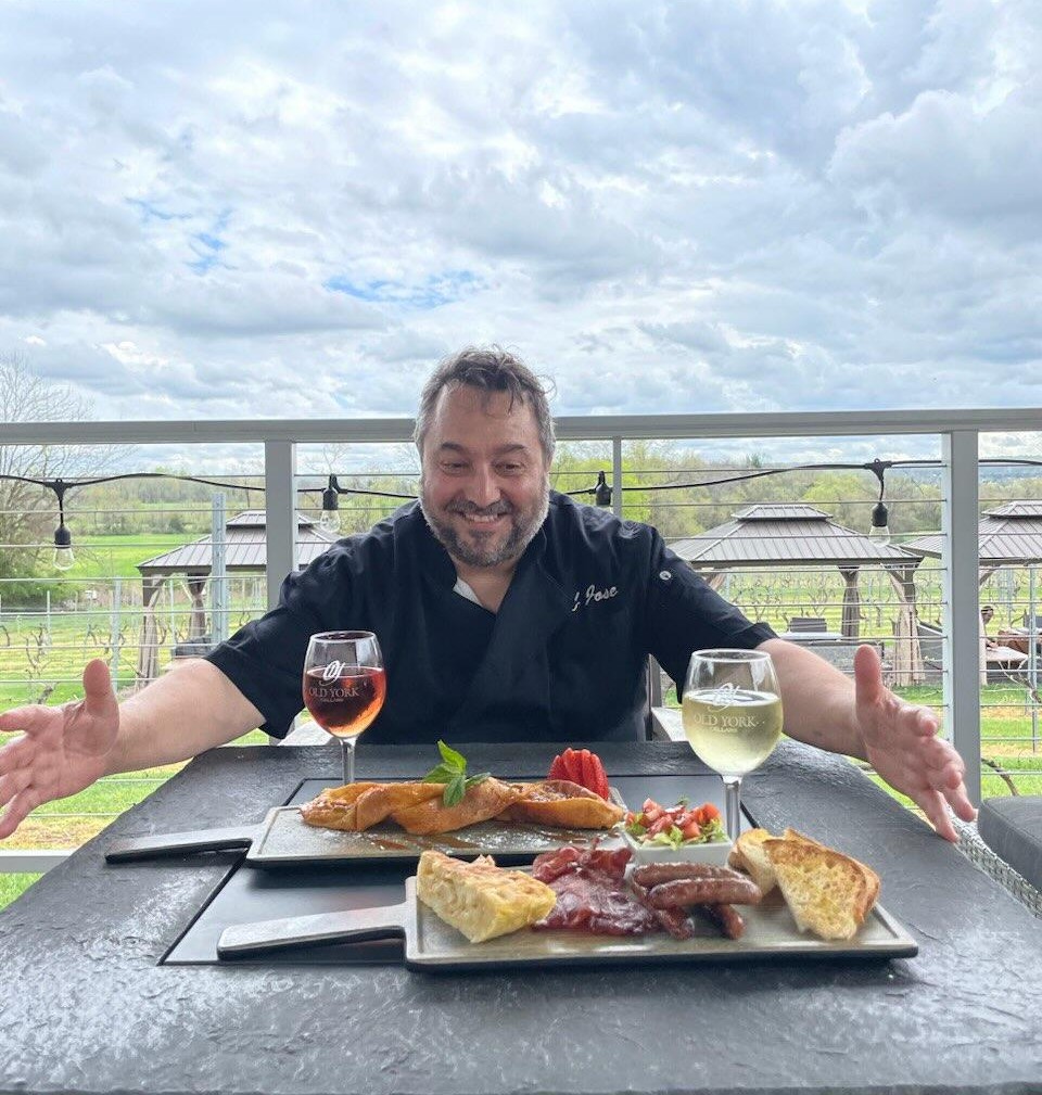 Chef José Diaz at Old York Cellars in Ringoes, NJ, smiling while presenting two plated dishes with glasses of rosé and white wine on an outdoor patio overlooking the vineyard.