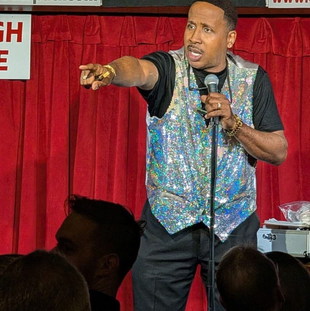 Comedian Derek Lee performing in front of a red curtain, pointing toward the audience while holding a microphone.  