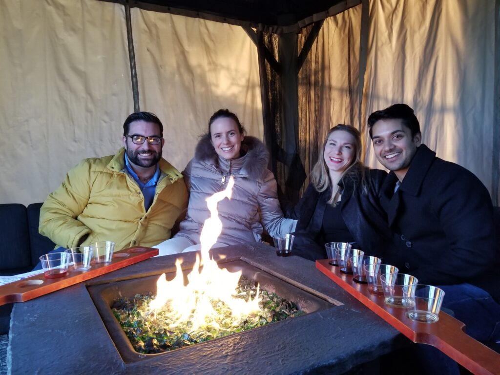 Group of four around a fire‑pit table enjoying wine and food pairings in a cozy outdoor setting.
