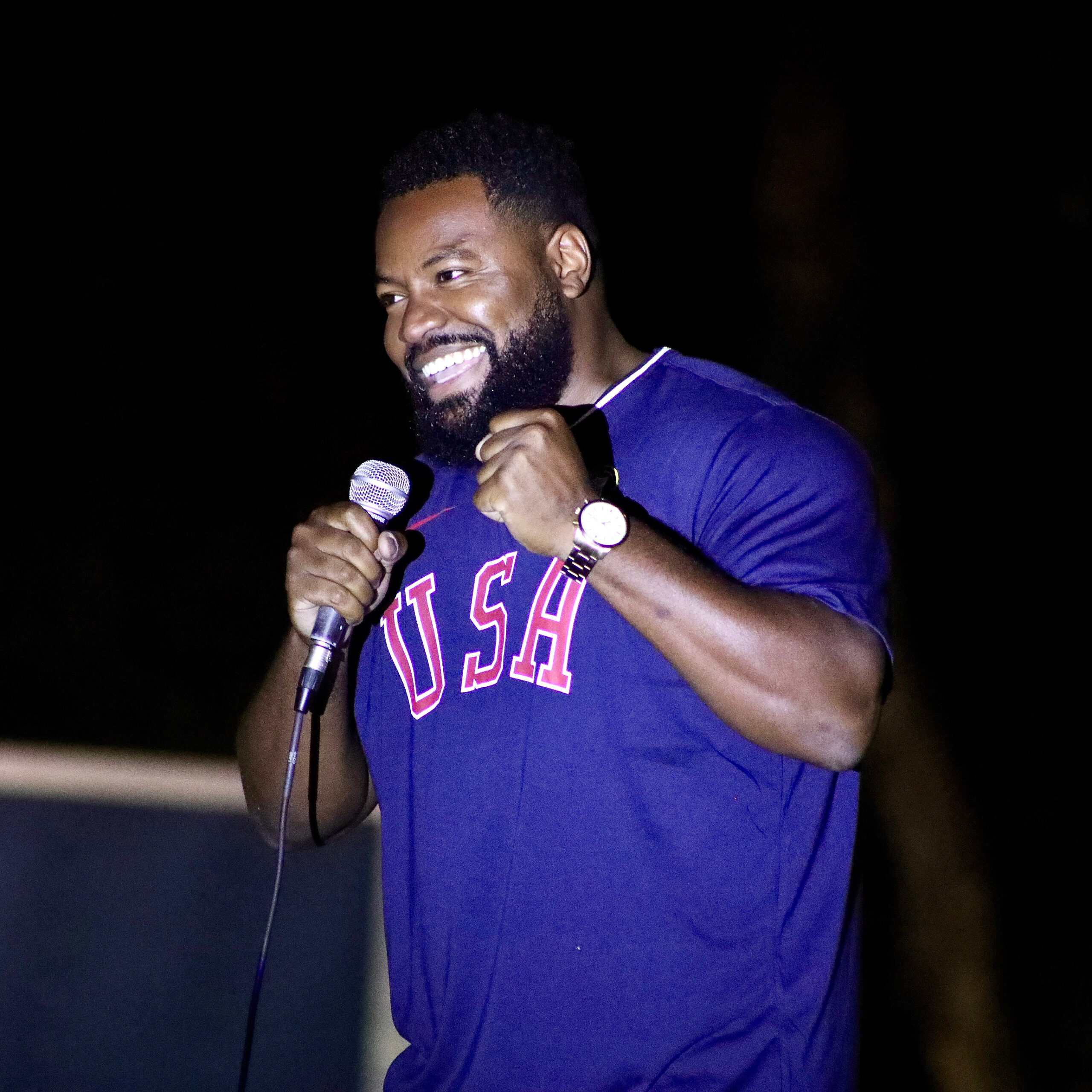 Comedian Gary Sharp Jr. smiling and holding a microphone while performing on stage, wearing a blue “USA” shirt.
