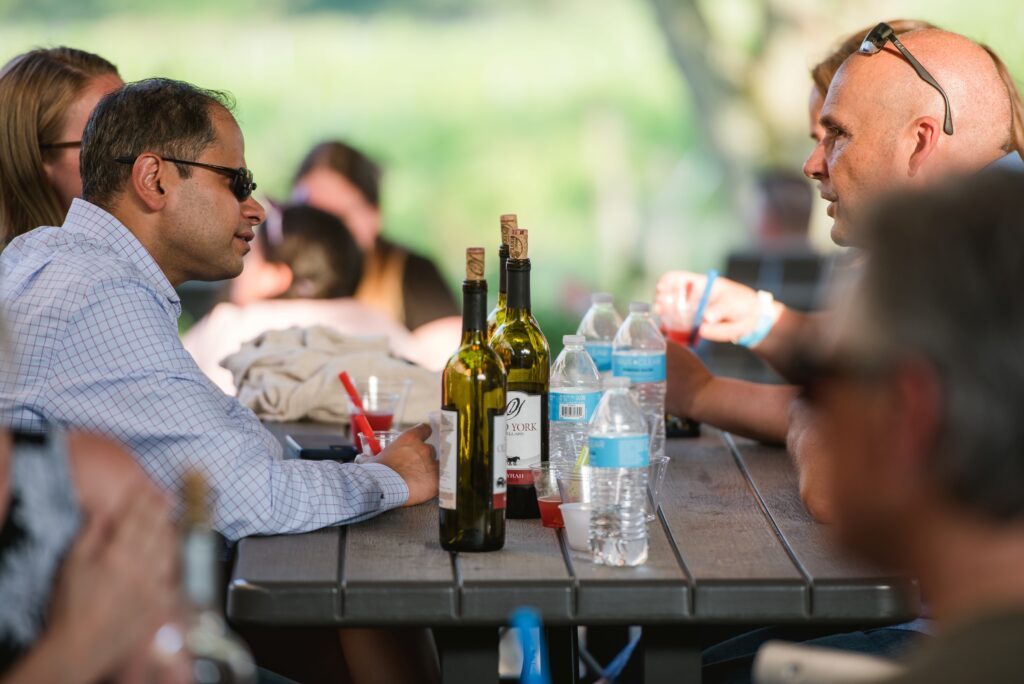 Friends sharing wine and conversation at a picnic‑style table at Old York Cellars in Ringoes, NJ, surrounded by greenery.
