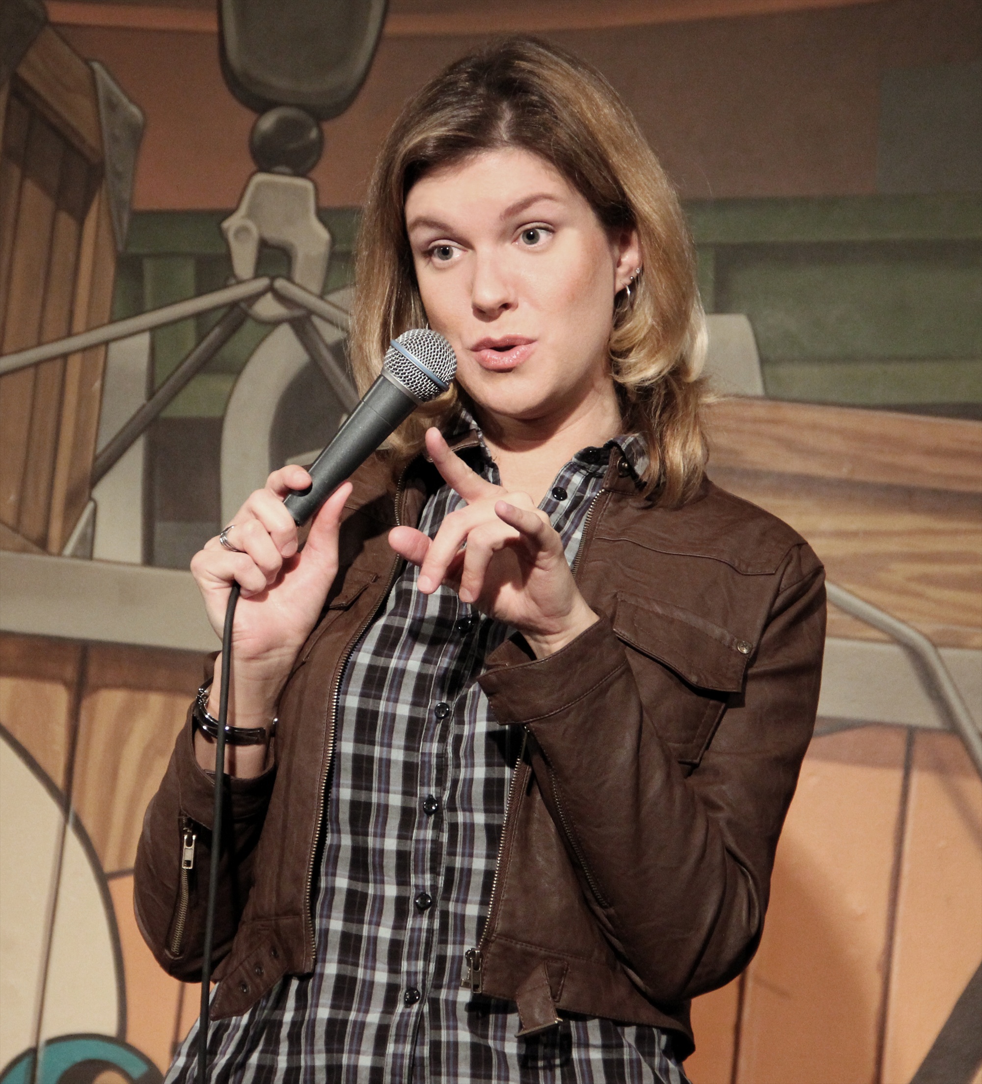 Comedian Kate Brindle gesturing with both hands while speaking into a microphone during a stand‑up set.