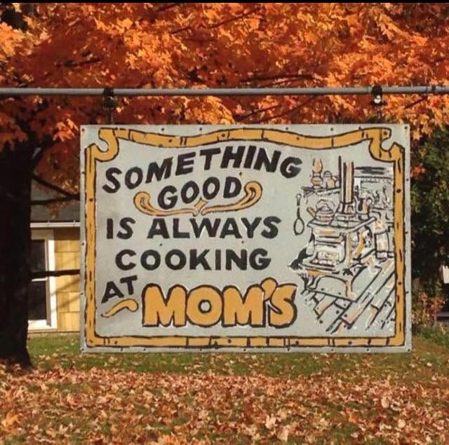 Roadside sign reading ‘Something Good Is Always Cooking at Mom’s,’ surrounded by autumn trees.