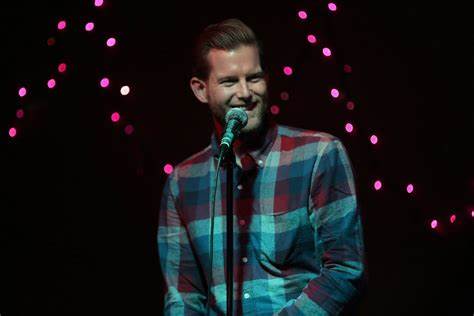 Comedian Patrick Holbert smiling and performing on stage in a blue and red plaid shirt, holding a microphone under soft stage lights.