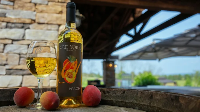 Bottle and glass of Old York Cellars Peach Wine displayed with fresh peaches grown at the winery on a wine barrel.