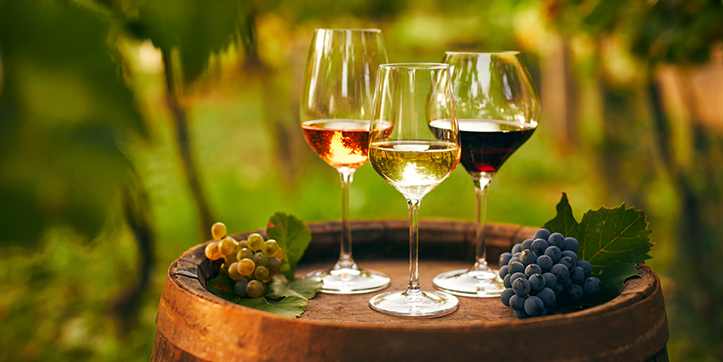 Red, white, and rosé wines in glasses beside grape clusters on an oak barrel in a vineyard.