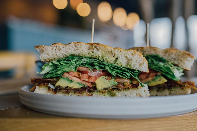 A fresh sandwich layered with tomato, avocado, and leafy greens on toasted bread, served on a white plate at Willow & Whisk restaurant that pairs meals with Old York Cellars wines.