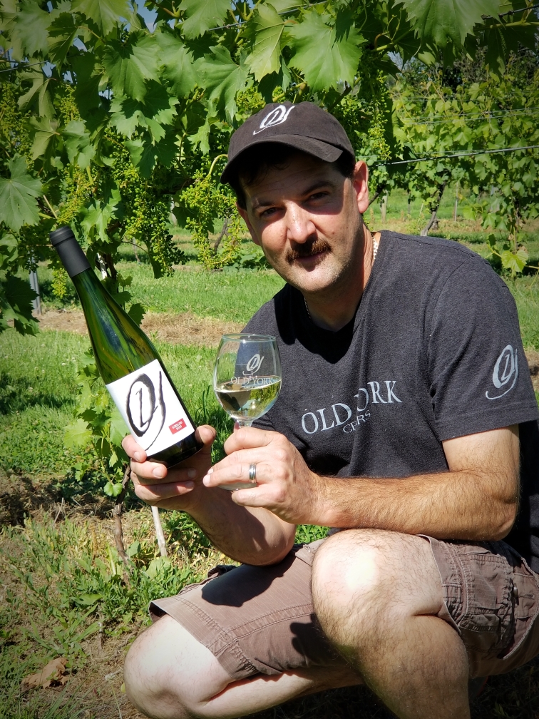 Winemaker, Scott Gares Winemaker kneeling among grapevines holding a bottle and glass of Old York Cellars Riesling white wine.