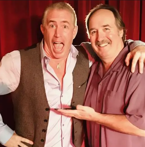 Comedians Scott Baker & Vinnie Mark posing together in front of a red curtain, smiling and gesturing playfully toward each other.