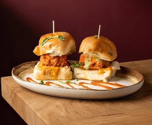 Two slider sandwiches with skewers on a plate drizzled with sauce, served on a wooden table.