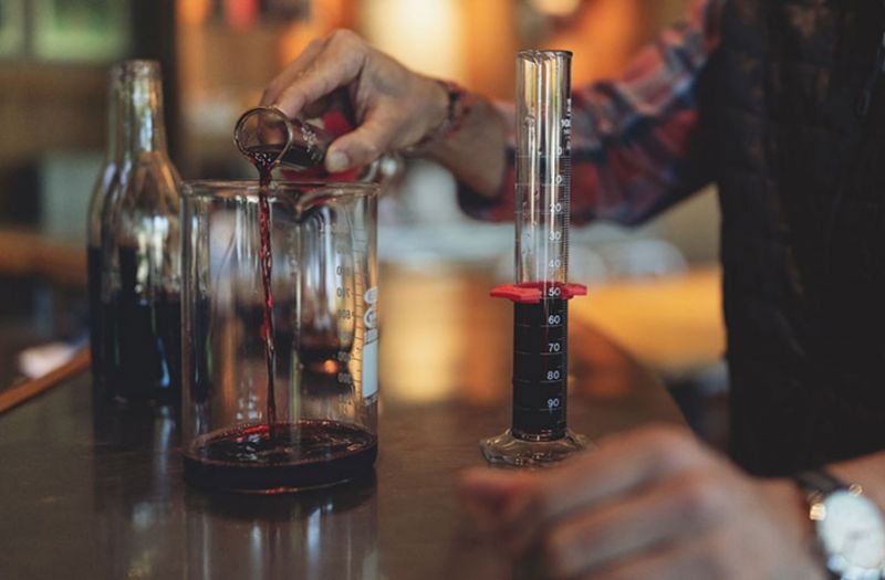 A Winemaker blending wines in glass cylinders and bottles on a table inside the winemaker's building.