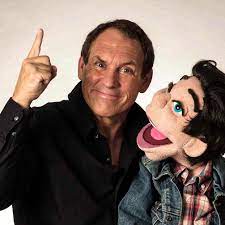 Comedian ventriloquist Taylor Mason smiling and gesturing beside a puppet with an expressive face.