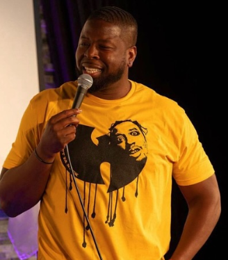 Comedian Tony Rogers performing on stage in a bright yellow T‑shirt with a graphic design, holding a microphone and smiling at the audience.
