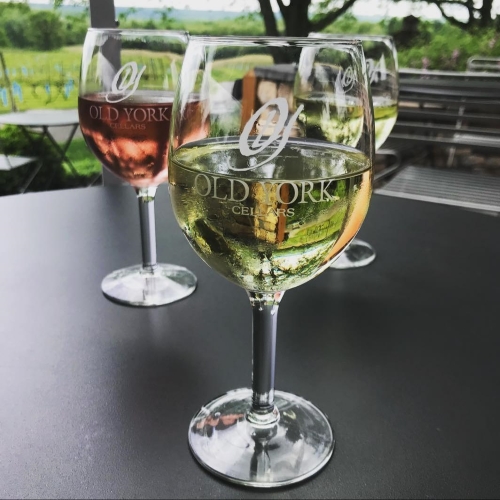 Glasses of Old York Cellars white and blush wines on an outdoor patio table overlooking green vineyard views.