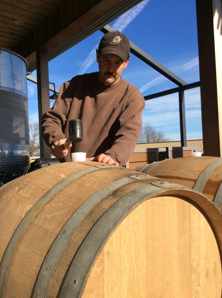 Winemaker, Scott Gares working on sealing an oak wine barrel at Old York Cellars Winery in Central NJ, under a covered area with tanks and a clear sky in the background.