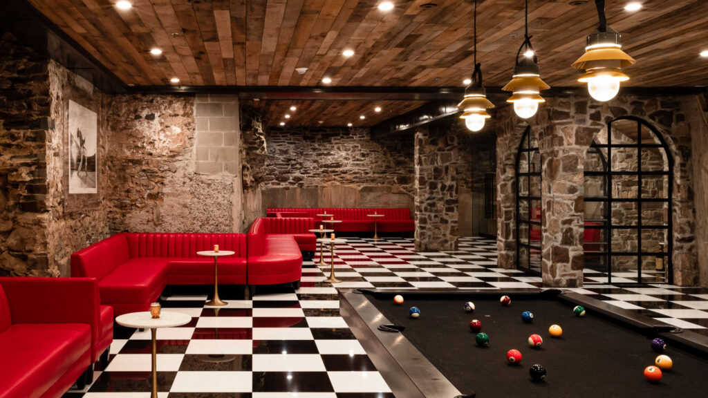 Recreation space at Logan Inn with a black‑and‑white checkered floor, red seating, stone walls, and a pool table under pendant lighting.