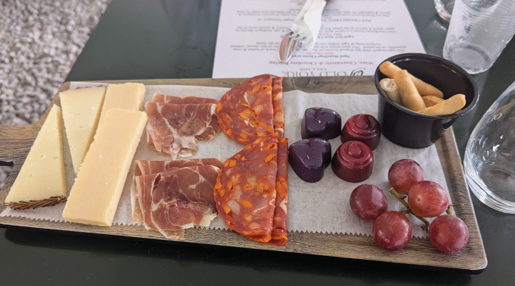 Charcuterie board at Old York Cellars in Ringoes, NJ featuring sliced cheeses, prosciutto, chorizo, artisan chocolates, red grapes, and crisp breadsticks. A tasting menu and glassware sit nearby, highlighting Central NJ wine and food pairing experiences.