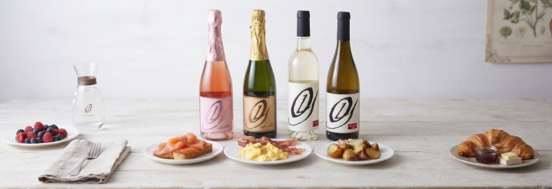 Four bottles of Old York Cellars wine—Sparkling Rosé, Sparkling White, Chardonnay, and Pinot Gris—displayed on a light wooden table with plates of brunch foods including smoked salmon, scrambled eggs, fruit, and a croissant, photographed at Old York Cellars Winery in Ringoes, NJ.