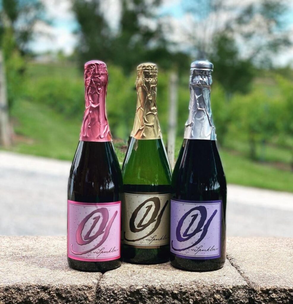 Three bottles of Old York Cellars sparkling wine—pink, gold, and silver labels—displayed outdoors at the winery’s vineyard in Hunterdon County, NJ.