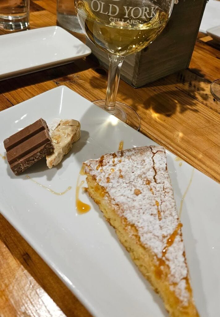 A slice of powdered‑sugar cake drizzled with caramel sits beside a piece of chocolate and nougat on a white plate, paired with a glass of white wine labeled “Old York Cellars.”