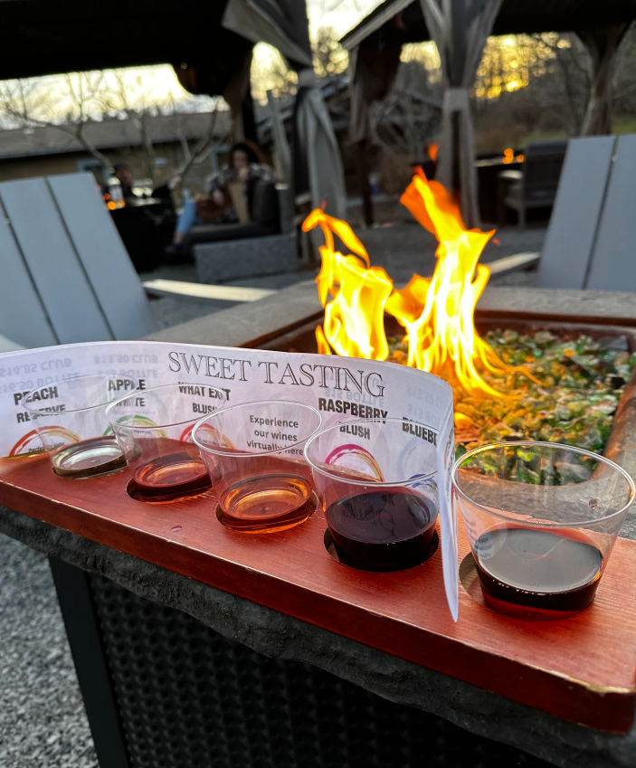 Sweet wine tasting at Old York Cellars in Ringoes, NJ. A tray of colorful wines labeled Peach, Apple, Vidal Blanc, Raspberry, and Blush sits beside a glowing fire pit in the cozy outdoor seating area.