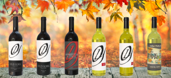 Six bottles of Old York Cellars wines—red and white varieties—lined up on a wooden surface with autumn leaves overhead.