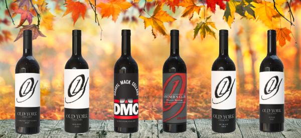 Six bottles of Old York Cellars red wines displayed on a wooden table with autumn foliage and warm golden background.